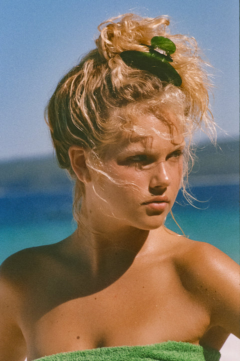 A close-up portrait of a woman outdoors by the water, with her hair in a messy bun and a green hair clip holding it back. She is wrapped in a green towel, with a blue sky and ocean in the background.